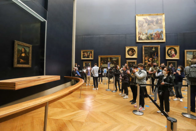 the Mona Lisa on the left hand side of the image behind glass, with people taking photos behind a barrier on the right. 