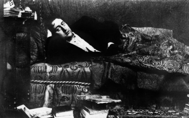 Black and white photo of Guillaume Apollinaire wearing a suit lying on a couch in his Paris apartment.