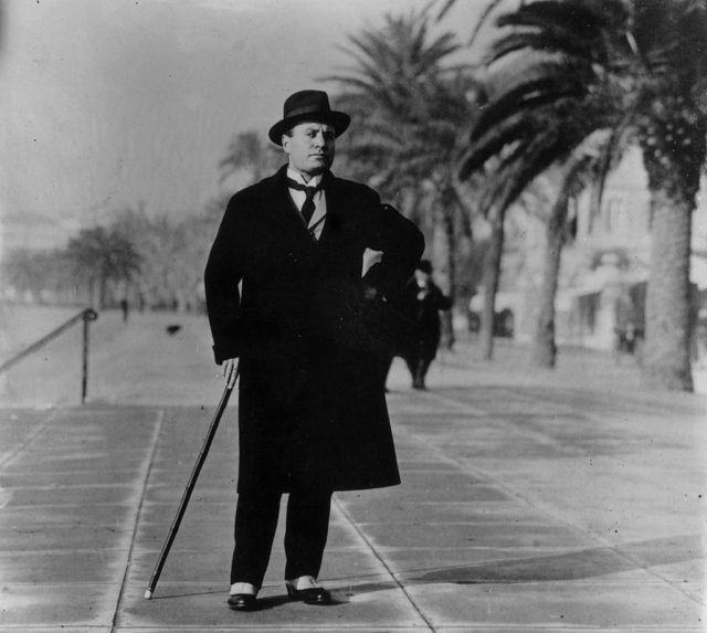 Black and white photo of Mussolini standing in a street with a cane. He is wearing all black with a coat and hat.