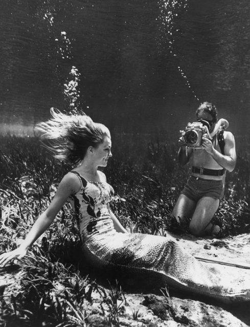 Black and white photo of a woman wearing a mermaid costume underwater while a photographer takes pictures of her.