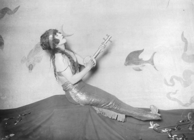 Black and white photo of a woman wearing a mermaid tail and holding a giant key sitting in front of a back drop painted with fish.