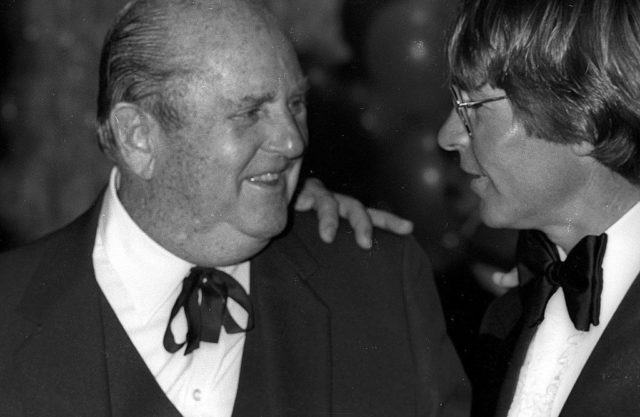 Black and white photo of Tom Parker, a balding man, in a suit, and John Denver, a man with a lot of hair and glasses, wearing a suit. 