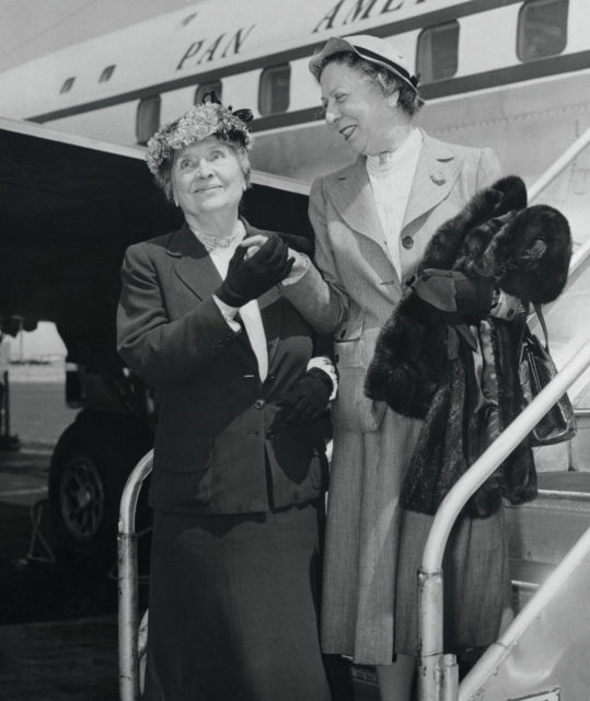 Helen Keller and Polly Thomson in skirts hold hands in from of plane