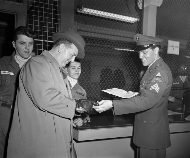 Black and white photo of Tom Parker in a fedora and a long coat and Elvis Presley in military uniform
