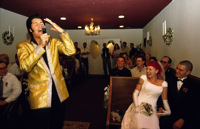 Man in gold sparkle vest singing into mic with wedding guests behind him in chapel