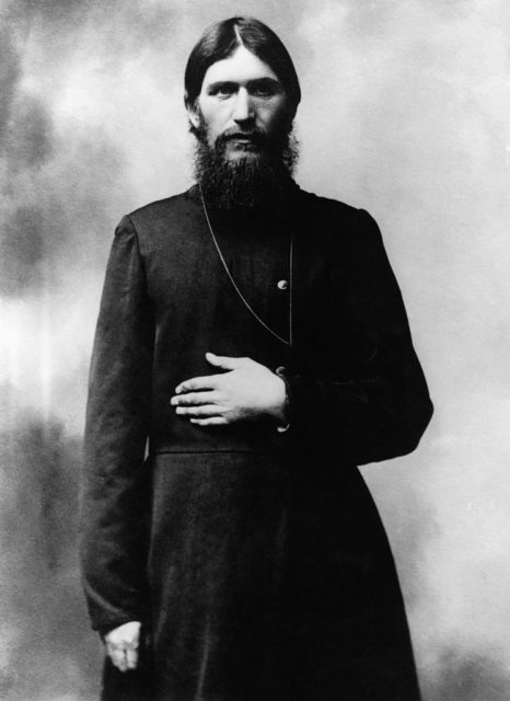 Black and white photo of Rasputin wearing a black robe standing up with one arm crossed over his stomach. Has his hair slicked down and a large black beard.