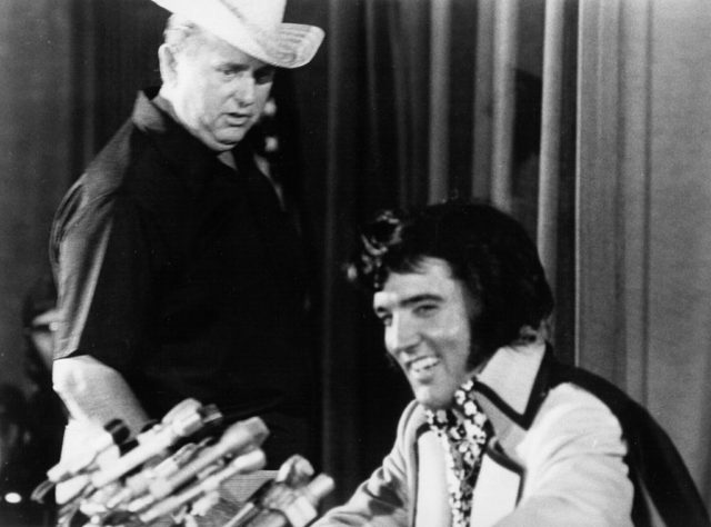 Black and white photo of Elvis sitting in front of microphones at a press conference with Tom Parker looking down over his shoulder while wearing a cowboy hat.