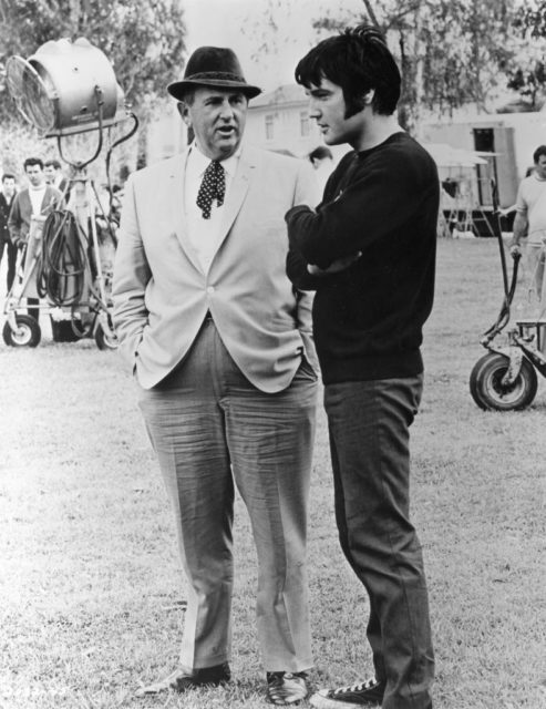 Black and white photo of Tom Parker in a light coloured suit and fedora standing beside Elvis, wearing a black shirt and dark pants.