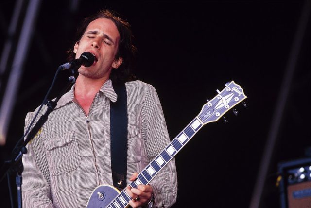 Jeff Buckley on stage singing and playing guitar.
