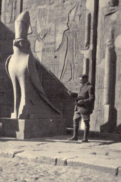 Black and white photo of man standing in front of large Egyptian bird statue and a wall with large engraved hieroglyphs 