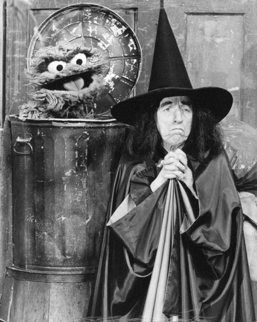 Black and white photo of the Wicked Witch of the West standing with her broom beside Oscar the Grouch in his garbage can.