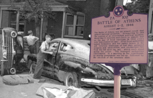 Black and white photo of a kid standing beside a car with a pink historical plaque in the foreground.