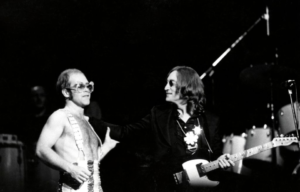 Black and white photo of a shirtless Elton John on stage with John Lennon who is holding a guitar wearing a blazer and sunglasses.