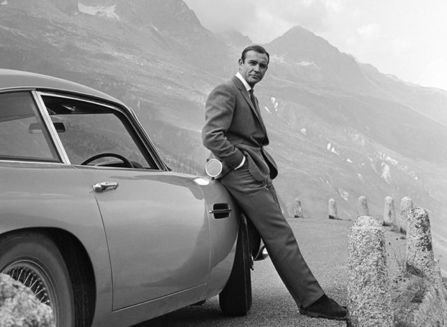 Black and white photo of Sean Connery in a suit leaning against an Aston Martin in front of mountains.