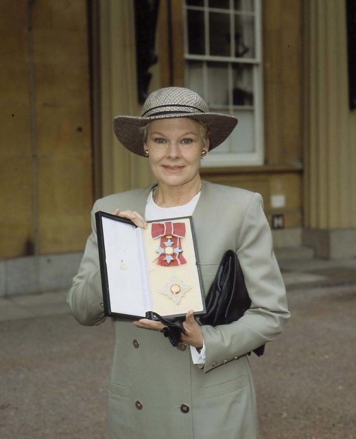 Colored photo of Judi Dench in a jacket and hat holding an award.