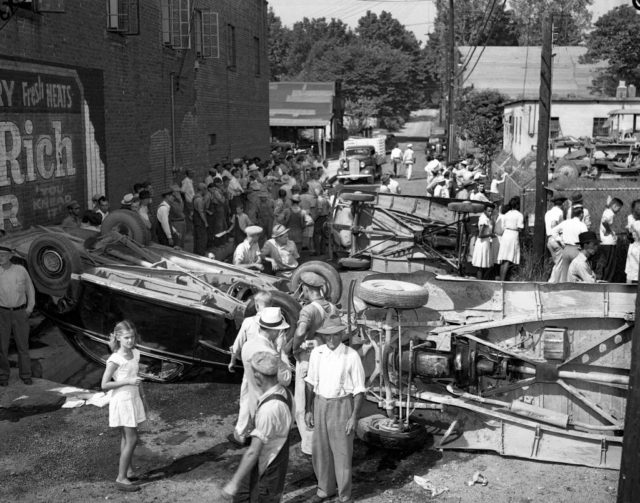 Black and white photo of a city street with many people walking around three overturned cars.