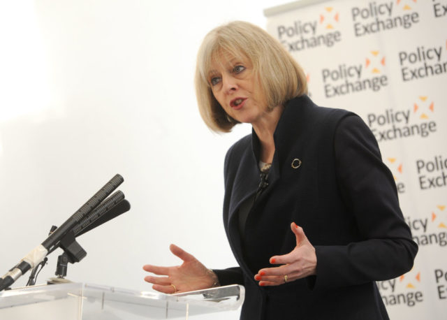 Colored photo of Theresa May giving a speech behind a podium, while wearing a black suit jacket.