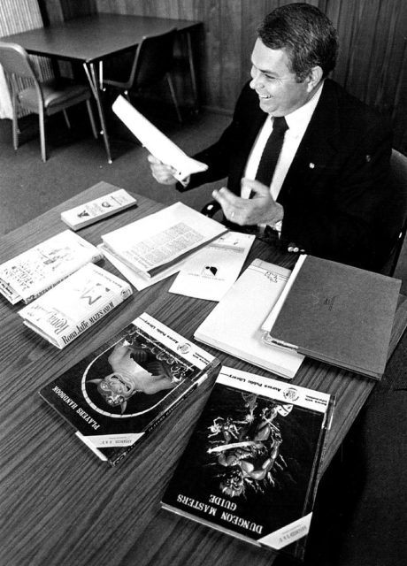 Black and white photo of Dave Price wearing a suit sitting at a table with Dungeons and Dragons books.
