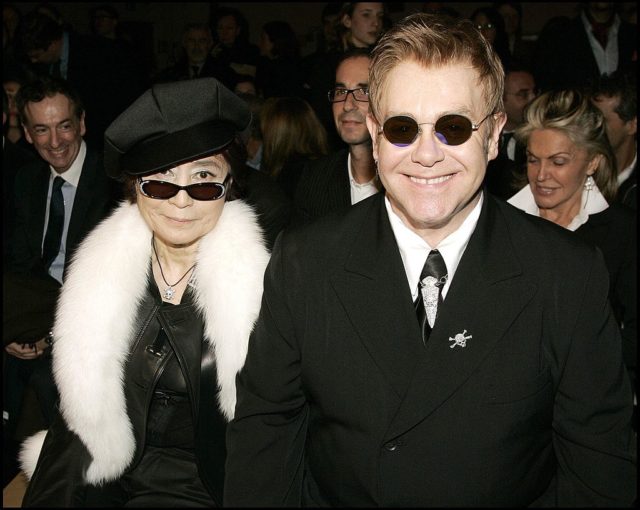 Colored photo of Elton John and Yoko Ono dressed in fancy clothes, Yoko in a dress, boa, sunglasses and a hat, Elton in a black suit and sunglasses.
