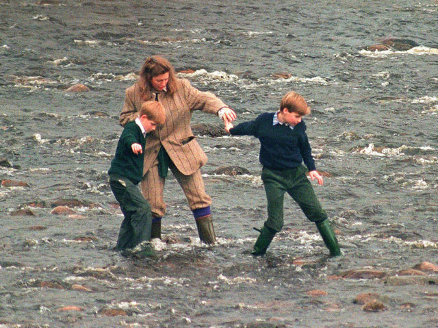 Colored photo of Tiggy Legge-Bourke in a tweed suit holding hands with a young Prince William and Harry. All three of them stand in the water.
