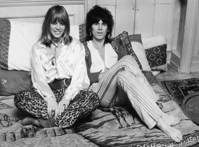 Black and white photo of Anita Pallenberg sitting cross legged and smiling beside keith Richards, with his legs crossed over each other.