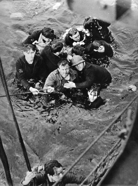 Black and white photo of men in military uniform floating in the water, hanging onto a rope off the side of a large ship.