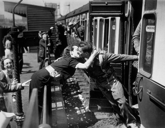 Black and white photo of a man in military uniform leaning out of a train to kiss a woman leaning over a railing.