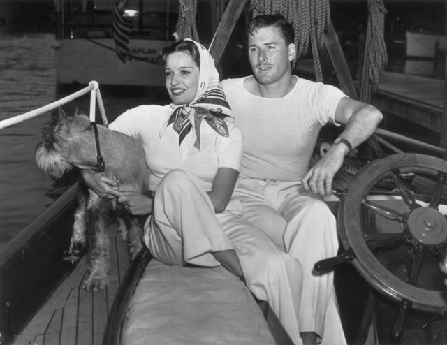 Errol Flynn holds the captain's wheel on board his yacht beside his wife Lili Damita and their dog. 