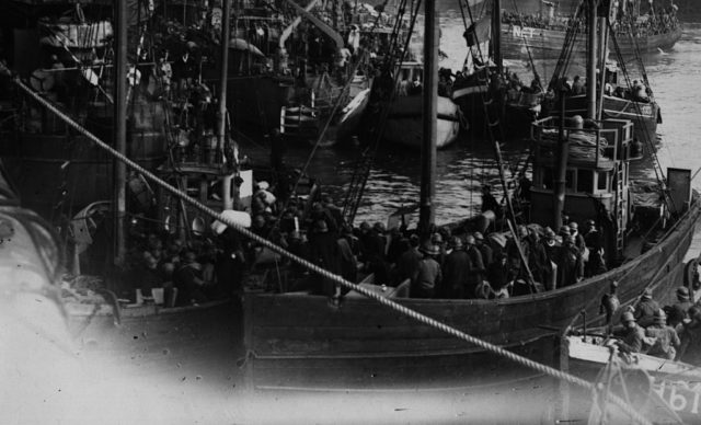 Black and white photo of several small sail boats tied together with their decks full of soldiers evacuating from Dunkirk.