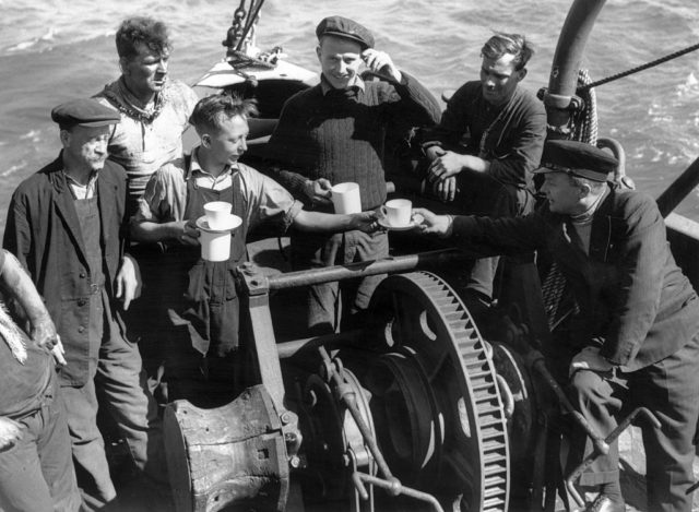Black and white photo of men in sweaters sitting at the back of a boat drinking cups of tea.