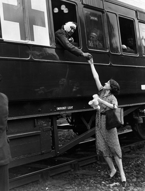 Black and white photo of a woman in a dress standing beside a train, handing something to a soldier learning out the window.