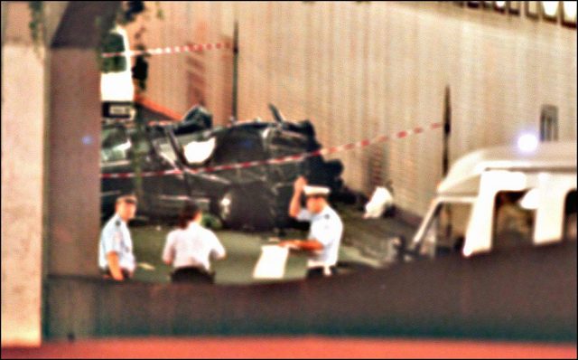 Colored photo taken from a distance of emergency personnel standing around a black car which has been squished from an accident.