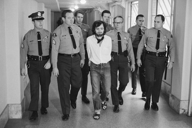 Black and white photo of Charles Manson being escorted by seven uniformed officers down a hallway.