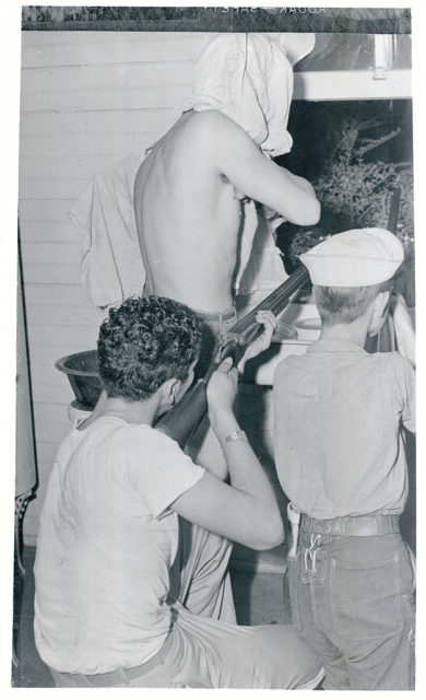 Black and white photo of three men with guns with their backs to the camera, aiming their guns out the window.