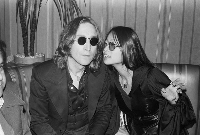 Black and white photo of John Lennon in a black suit and sunglasses beside May Pang, wearing a black leather vest over a black shirt and sunglasses. May is whispering something to John.