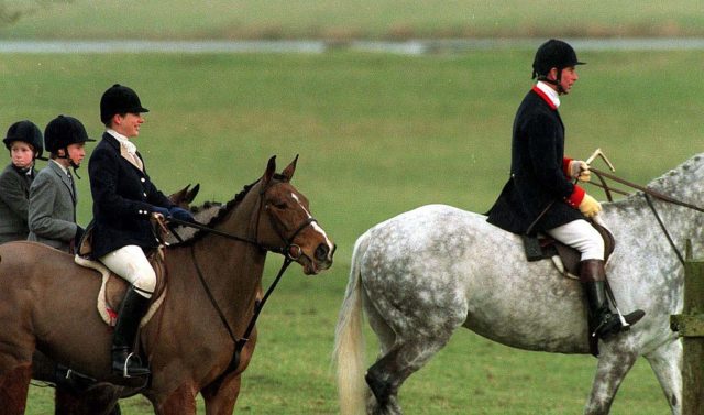Colored photo of Prince Charles on a grey horse, Tiggy Legge-Bourke on a brown horse, and Prince William and Prince Harry's heads popping up behind her on their own horses.
