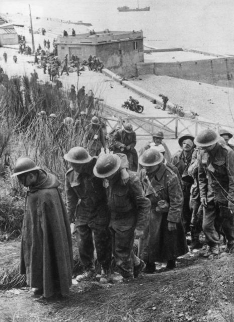 Black and white photo of men in military uniforms and tin fats walking up a hill away from a beach.