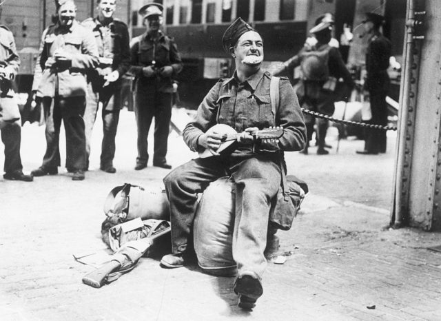 Black and white photo of a man in military uniform sitting on a bag playing a banjo and eating a Cornish pasty. 