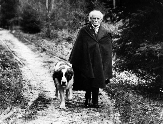 Black and white photo of David Lloyd George wearing a black cloak walking his dog.