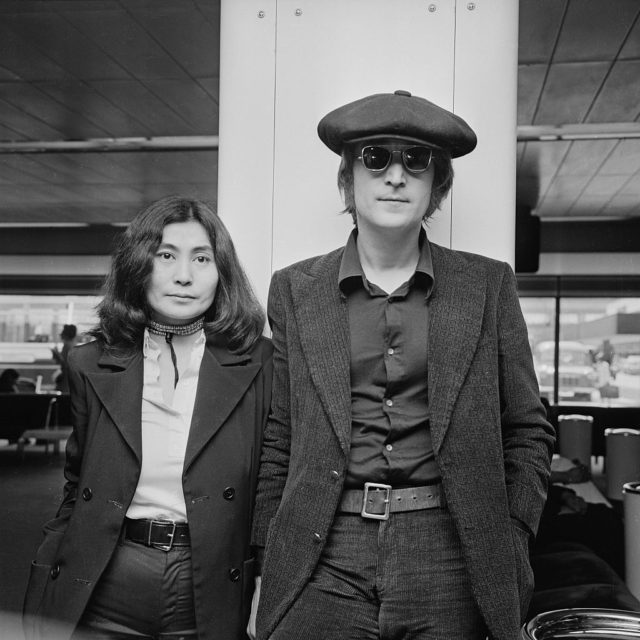Black and white photo of John Lennon and Yoko Ono both wearing blazers and pants, Lennon with a flat cap and sunglasses.