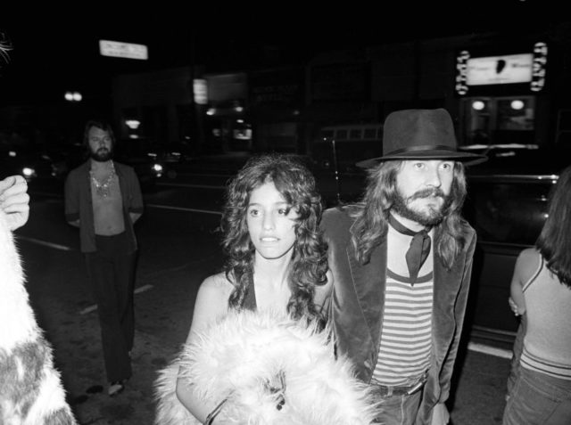 Black and white photo of Lori Maddox holding a feather boa beside John Bonham wearing a hat and striped shirt.
