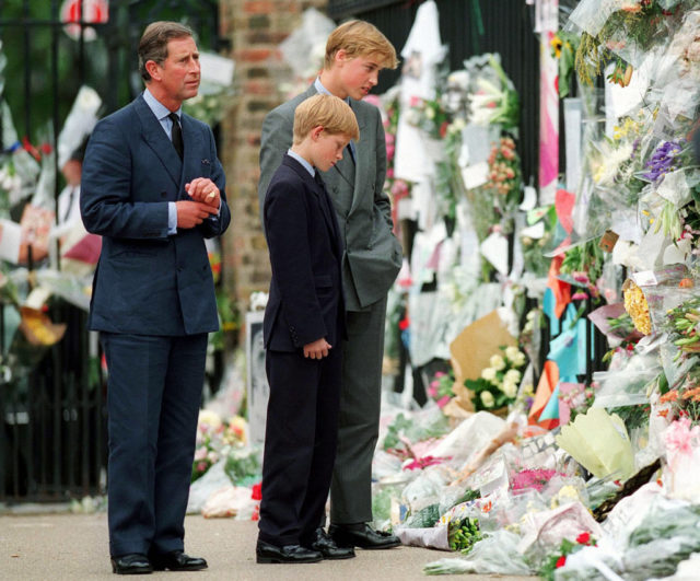 Colored photo of Prince Charles, Prince William, and Prince Harry wearing suits and looking at a large number of flowers placed in honor of Diana.