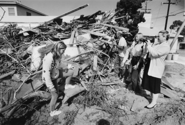Black and white photo of a pile of rubble from the destroyed McMartin Preschool building with people standing around, and one woman filming.