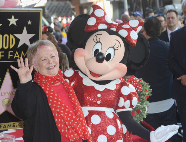 Colored photo of Russi Taylor waving at the camera while standing beside Minnie Mouse wearing her red and white polka dot dress and bow.