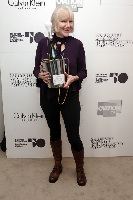 Colored photo of Cynthia Albritton with short blonde hair smiling at the camera while holding a bucket with champagne. 