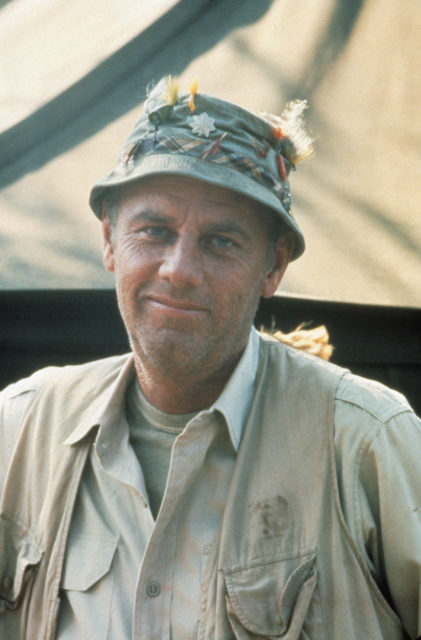 Colored photo of McLean Stevenson as Henry Blake wearing a fishing hat and tan vest.