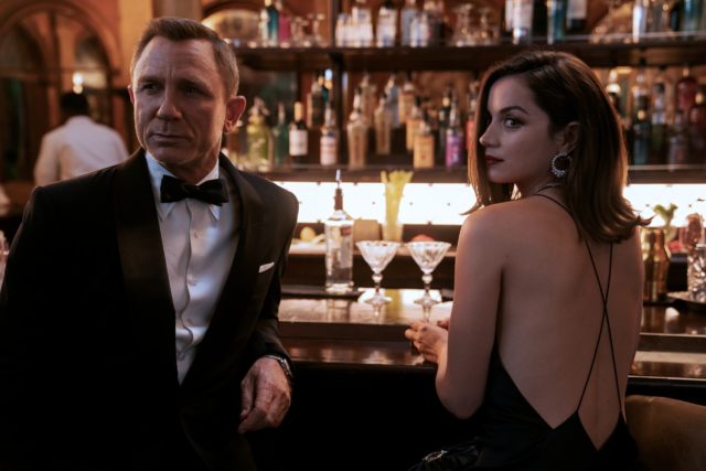 Colored movie still of Daniel Craig and Ana De Armas in No TIme To Die, sitting at a bar while wearing fancy dress.