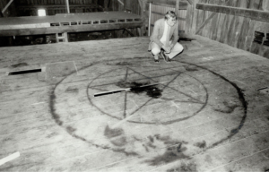 Black and white photo of a man in a suit kneeling down looking at a pentagram painted on a wooden barn floor.