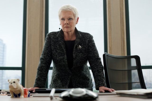 Colored movie still of Judi Dench in Skyfall standing up over a desk with her hands resting on it.