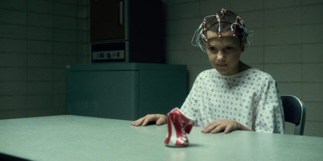 Colored photo of Millie Bobby Brown as Eleven in a hospital gown with shaved hair and electrodes on her head, crushing a can of coke.